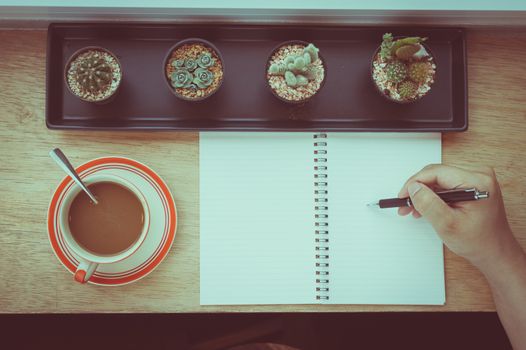 Top view of male hand writing on notebook and coffee cup on wood bar beside window in cafe in morning time with film filter effect