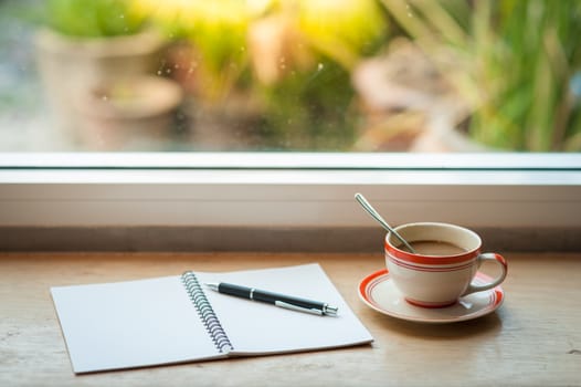 Coffee cup and opened notebook with pen on wood bar beside window at coffee shop in morning time