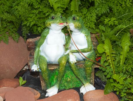 Two green ornament frog sitting on a bench near a stones in the tall grass