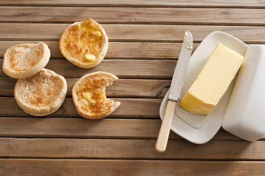 Freshly baked and toasted tasty crumpets, one bitten into, on a wooden table with a pat of butter on a plate, overhead view