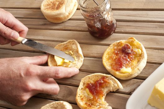 Man spreading freshly baked and toasted crumpets with jam and butter on a wooden slatted table, overhead view
