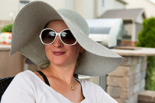 Head and Shoulders Close Up of Joyful Woman Wearing Large Brimmed Sun Hat and Sunglasses Smiling and Looking to the Side Outdoors on Backyard Patio on Sunny Day