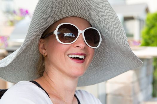Head and Shoulders Close Up of Joyful Woman Wearing Large Brimmed Sun Hat and Sunglasses Laughing Outdoors on Backyard Patio on Sunny Day