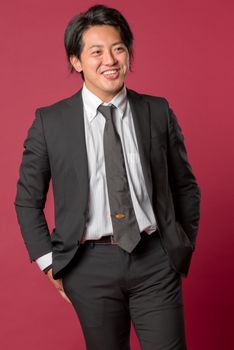 A half body portrait of a young Japanese man in a business suit laughing on a red background.