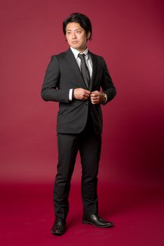 A portrait of a young Japanese man in a business suit standing in a strong pose while buttoning his Jacket on a red background.