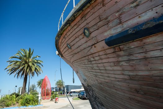 View of bow of an old wooden boat in dry dock. Small port holes can be seen along with palm trees in background.