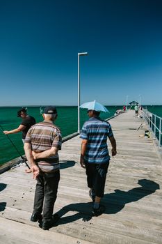 MELBOURNE/AUSTRALIA - FEBRUARY 6: Locals fishing on the jetty in Mordialloc which is a coastal suburb of Melbourne, Ausralia. In the foreground a man walks with his friend who is wearing an unbrella for a hat.