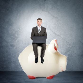 Businessman working on laptop and sitting  on piggy bank on grey wall background