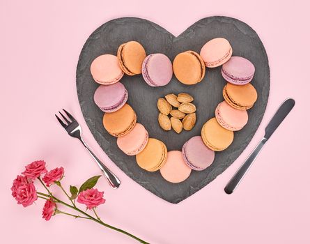 Still life, macarons sweet colorful, heart shape, fork knife. Black placemat, roses almond. French dessert, table setting. Unusual creative romantic, pink background. Concept love story.Valentines Day