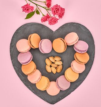 Still life, macarons sweet colorful, heart shape, black placemat. French traditional dessert, roses, almond. Unusual creative romantic, pink background. Concept for love story. Valentines Day