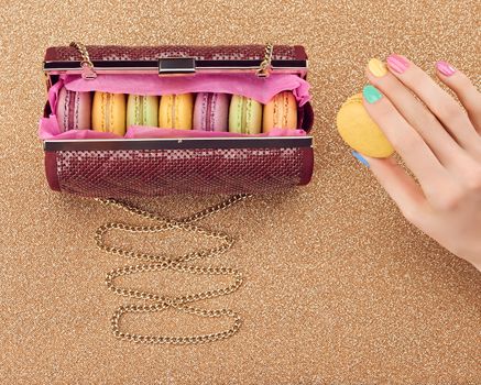 Macarons french in handbag, woman hand. Luxury shiny glamor fashion clutch. Sweet colorful dessert. Unusual creative art, gold party background, bokeh, copyspase. Vintage