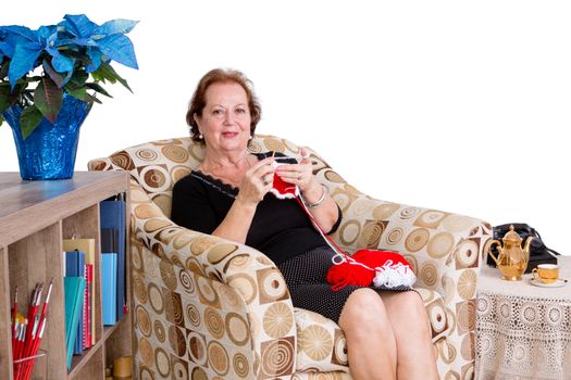 Elegant elderly lady sitting knitting at home in a comfortable armchair looking up to give the camera a friendly smile against a white wall with copyspace