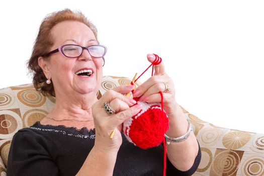 Happy elderly lady enjoying a joke laughing as she concentrates on her colorful festive red knitting while relaxing in a comfortable armchair, isolated on white