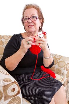 Senior lady in a stylish black dress wearing eyeglasses relaxing with her knitting in a comfortable armchair as she smiles at the camera, isolated on white