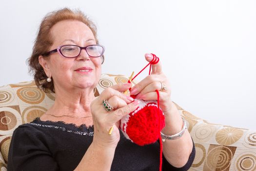 Senior lady sitting knitting at home holding up her colorful red knitting in front of her with a pleased look of concentration, isolated on white