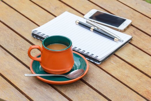 Hot coffee and notebook and pens and smart phone on the wooden table. Focus on a cup.