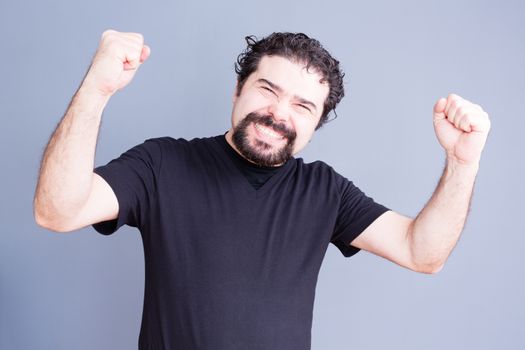 Single handsome bearded man in black shirt holding fists up with elated expression over gray background