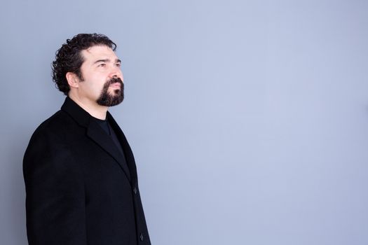 Three quarter view of hopeful attractive dark haired and bearded middle aged male in black shirt and blazer looking away over gray background with copy space