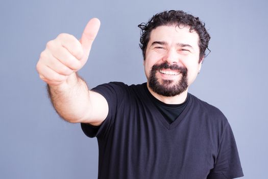 Middle aged bearded single man in black shirt sticking his hand outward with thumb up and big happy smile over gray background