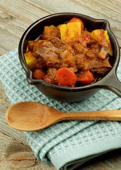 Delicious Beef Stew with Carrots, Potatoes, Celery and Leek in Cast Iron with Napkin and Wooden Spoon closeup on Wooden background
