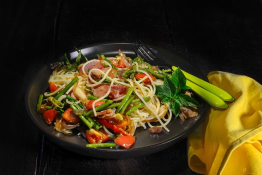 Spaghetti with asparagus tomatoes bacon and herbs on wood table in a black plate
