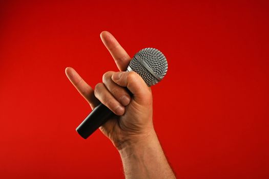 Man hand holding voice microphone with devil horns rock metal gesture sign over red background, diagonal