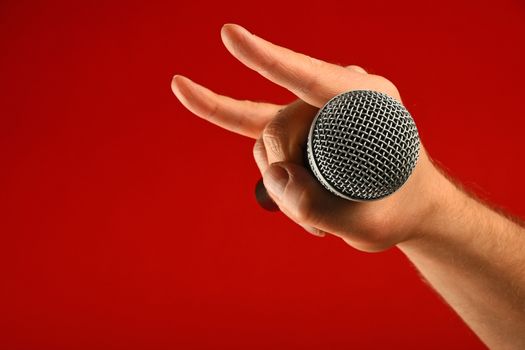 Man hand holding voice microphone with devil horns rock metal gesture sign over red background, side view