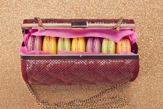 Macarons french in handbag. Luxury shiny glamor fashion clutch. Sweet colorful dessert. Unusual creative art, gold party background, bokeh, closeup. Vintage