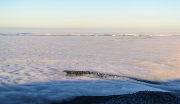 View from the top of the mountain above the clouds. Scenic landscape of clouds over the land. The sky is clear. Evening sunlight falling on the clouds. Some mountains are seen on the horizon. Winter