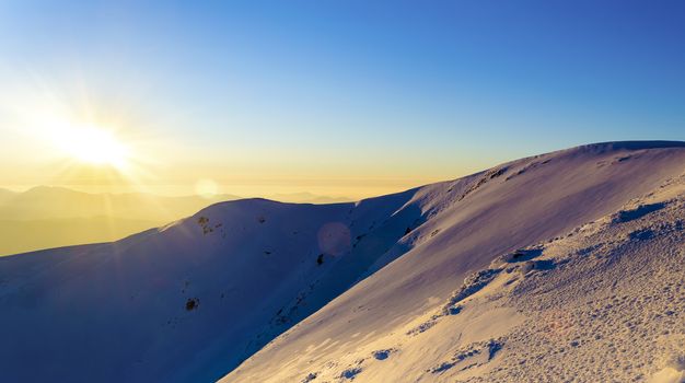 Sunset in the winter snowy mountains. Direct sunlight. Sun beam. Light reflected from the snow. Clear blue sky. Deep shadows