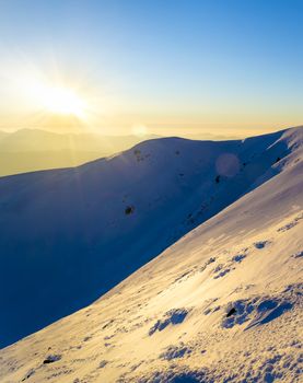 Winter sunset in the mountains. Direct sunlight. Sun beam. Light reflected from the snow. Steep slope. Clear blue sky. Deep shadows. Ukraine