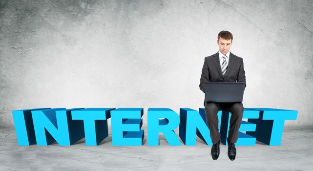 Businessman working on laptop and sitting on word internet on grey wall background