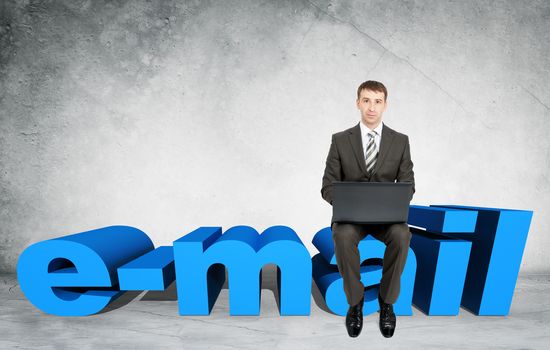 Businessman working on laptop and sitting on word e-mail on grey wall background