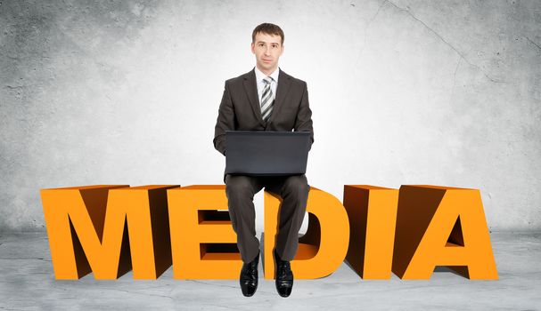 Businessman working on laptop and sitting on word media on grey wall background