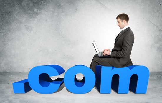 Businessman working on laptop and sitting on word dot com on grey wall background