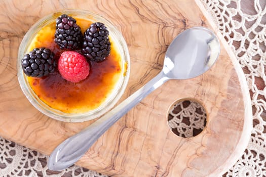 Scrumptious creme brulee topped with red and black raspberries from point of view angle with spoon over doily and wooden tray