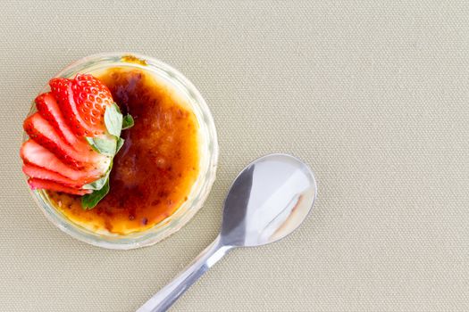 Delicious custard dessert topped with strawberry slices from point of view angle with spoon over brown tablecloth