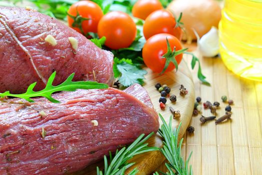 Raw marinated beef on bamboo cutting Board beside the tomatoes, rosemary, arugula, pepper, cloves, garlic and vegetable oil.