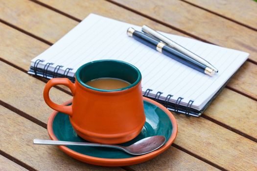 Hot coffee and notebook and pens on the wooden table. Focus on a cup.