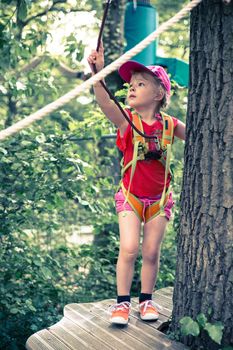 Little blond girl standing on a wooden platform doing a ropes course in an adventure park