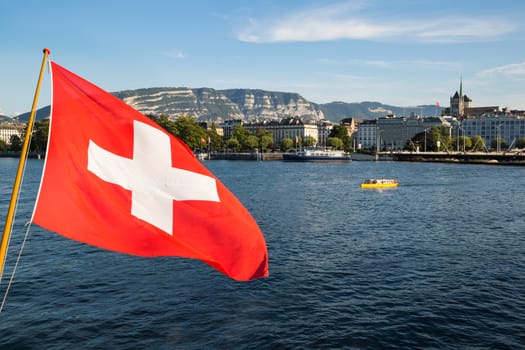 A swiss flag floating in the wind over the Lake Geneva, with the city of Geneva in the background.