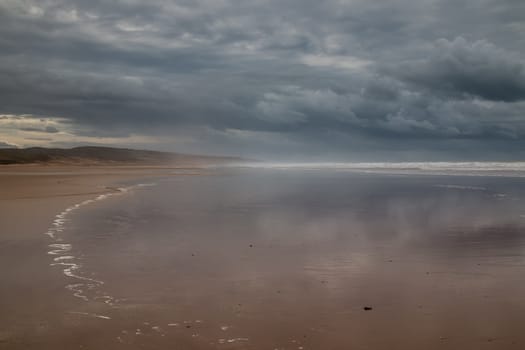 Stormy weather with heavy clouds. Beach with a wavy water of the Atlantic ocean during a tide. Mountain in the background.