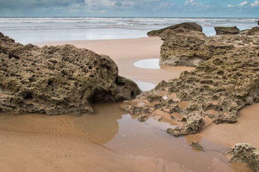 Rocks on the sandy beach in the foreground. Windy autumn day with many clouds. Waters of Atlantic Ocean in the background. Moulay Bouzerktoun, Morocco.