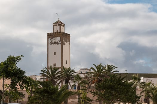 Minaret of an old mosque in Essaouira, among the palms of the city park. Cloudy sky.