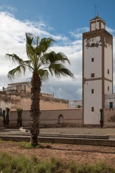 Minaret of an old mosque in Essaouira and a palm tree. Cloudy sky.