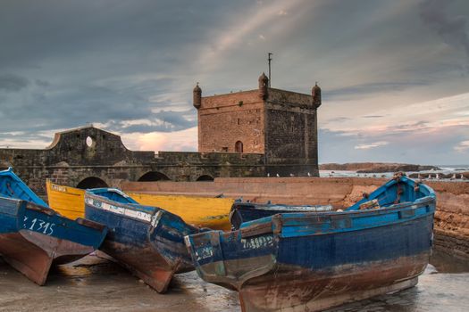 Parked boats in the port of Essaouira. Tower as a part of city fortification in the background. Early morning cloudy sky.