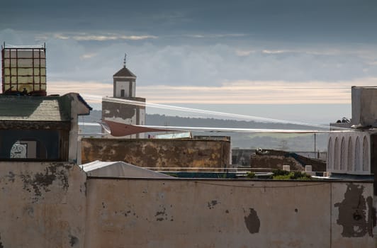 View from a roof on another roofs and minaret of a mosque. Hill with trees in the background. Cloudy sky.