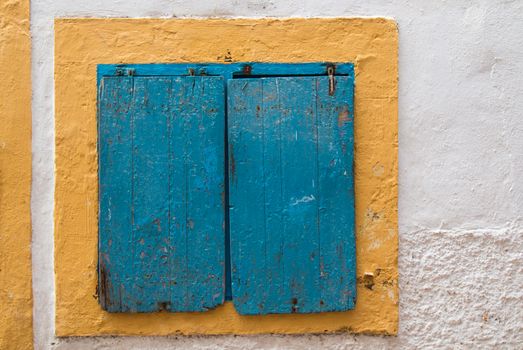 White wall with a window covered by blue shutter. Yellow frame of the window.