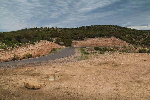 Moroccan country. Dry soil in the foreground with a road and argan trees garden on a hill. Cloudy sky.