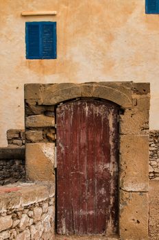 Old wooden gate in a stone frame. Orange facade and windows with blue shutters. Essaouira, Morocco.